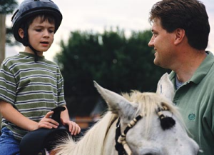 Young boy wearing a riding helmet sits on a horse being held by his father.