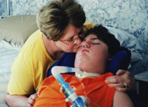 Mother kissing a young man with significant disabilities in the bedroom of a home.