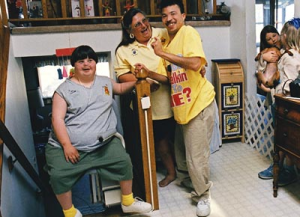 Woman laughs with a young man and a young woman with disabilities seated on a lift at the top of a flight of stairs in a home.
